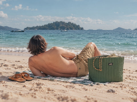 Sonny relaxing on a beach towel beside his OriGinaLe Millie Carry-All tote bag in green, overlooking the tropical sea.